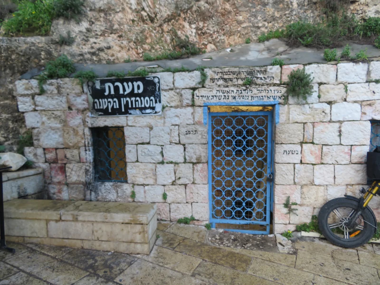 חזית מערת הסנהדרין הקטנה: קיר אבן, שלט ושער ברזל כחול בפתח