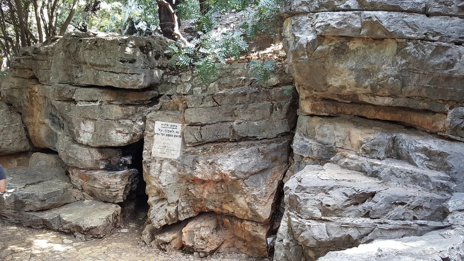 פתח מערת רשב"י בין סלעי הגיר המשוכבים בפקיעין, ולצדו שלט האתר