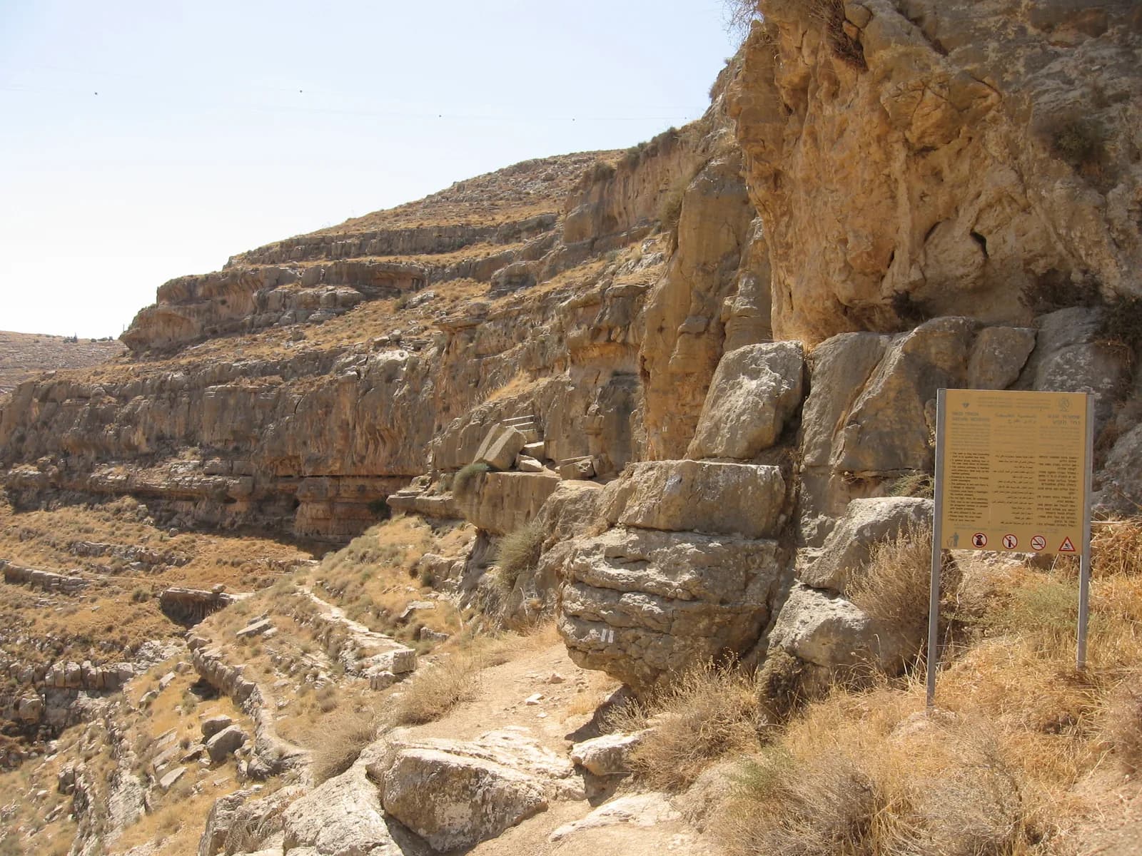 השביל על מדרגת הסלע אל פתח מערת חריטון, עם גושי הסלע ושלט ההסבר, מעל קניון נחל תקוע