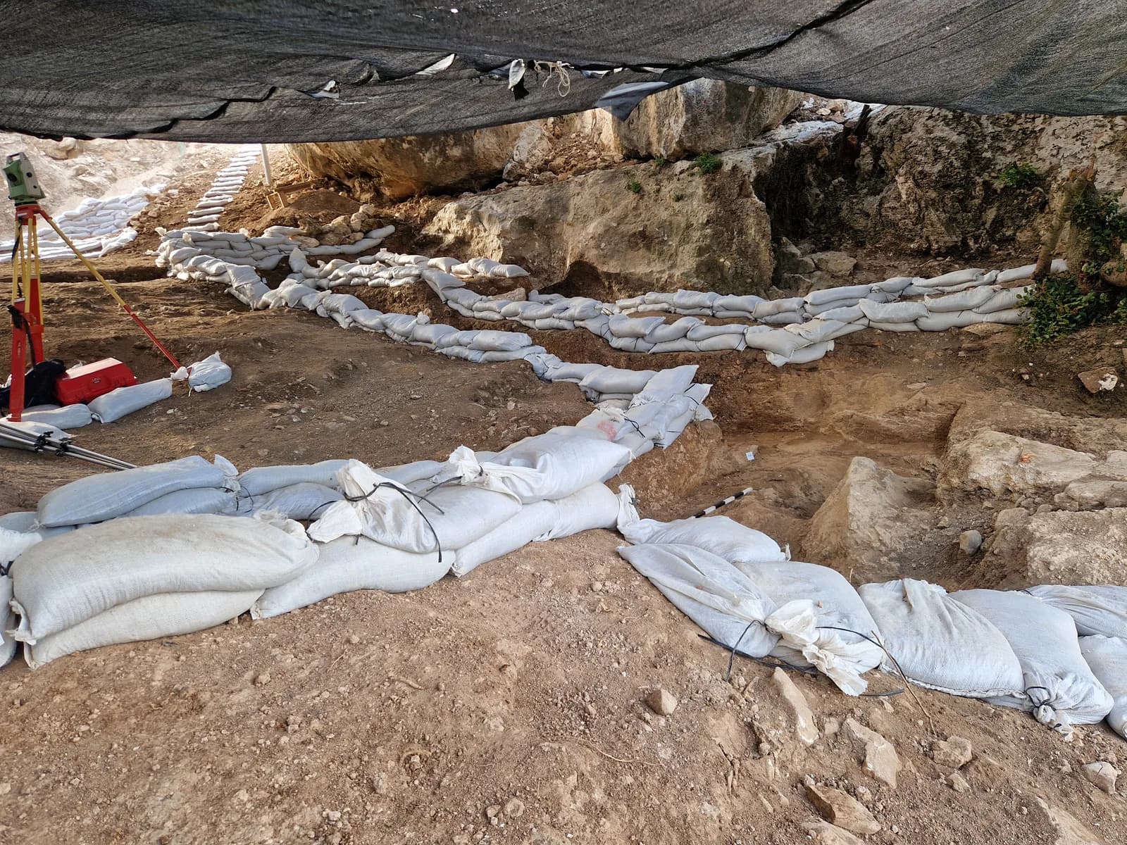 שטח החפירה במערה תחת יריעות צל, עם שקי חול לאורך חתכי החפירה