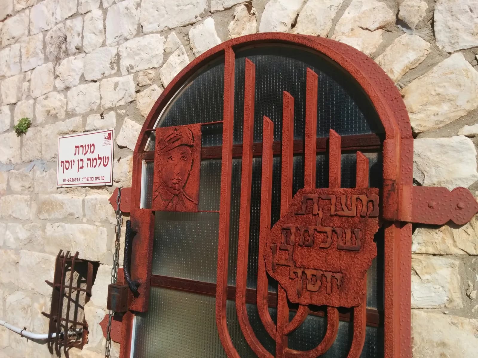 דלת הברזל המעוטרת בכניסה למערת שלמה בן יוסף, ובה דיוקנו ומנורה, ולצדה שלט האתר